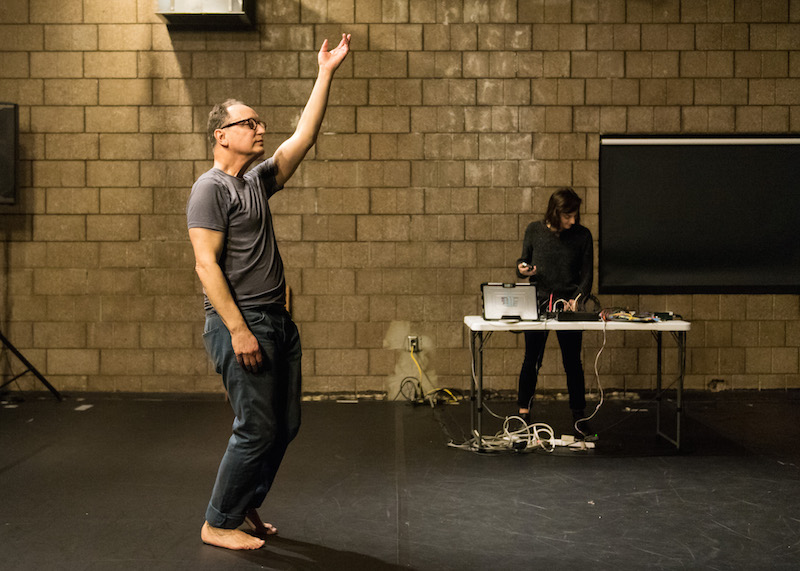 Paul Lazar in jeans and a t-shirt stands with his left arm extended to the ceiling